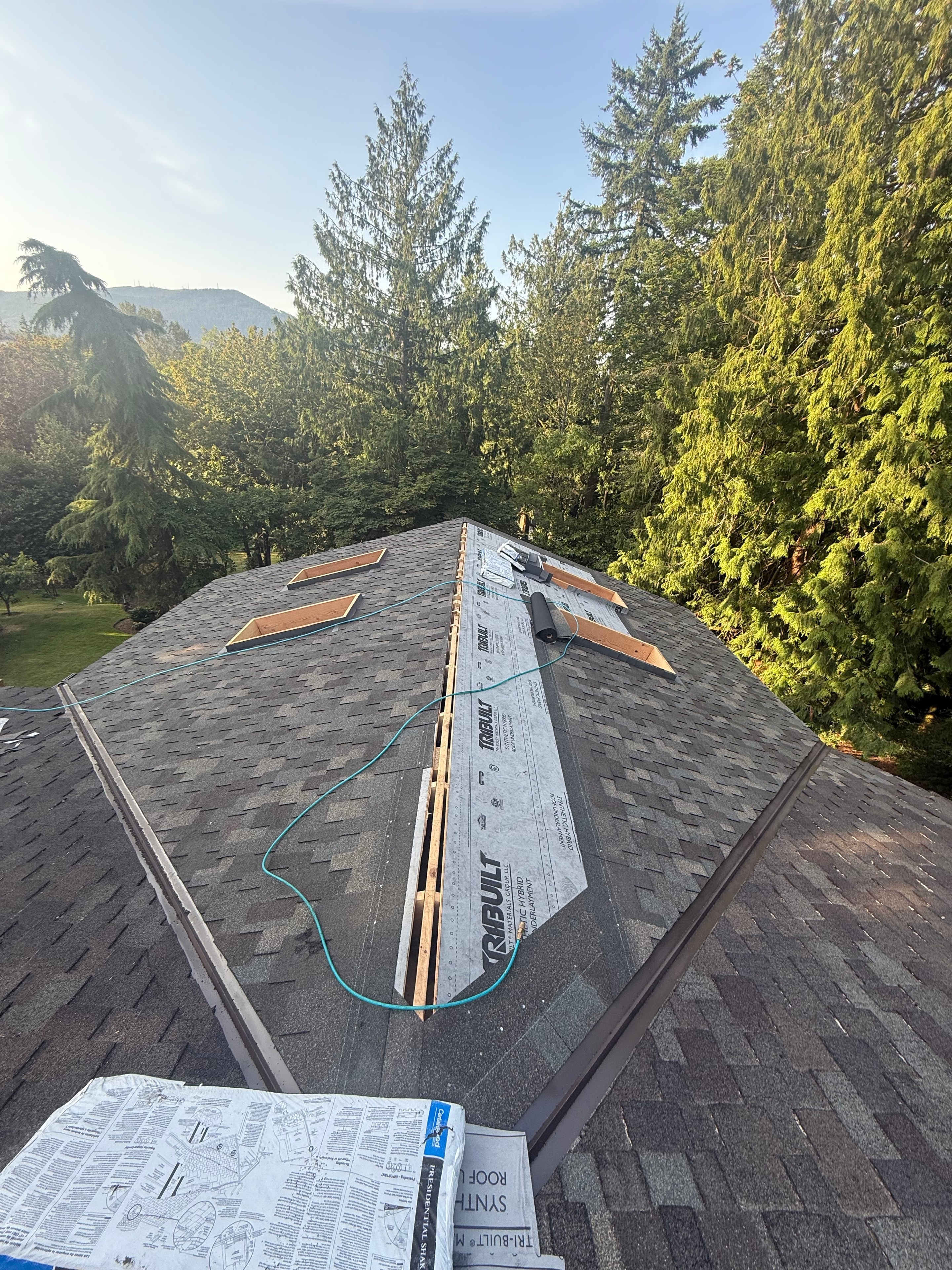 Wide aerial view of residential roof with multiple skylight openings framed, TRI-BUILT membrane on ridge, mountain backdrop